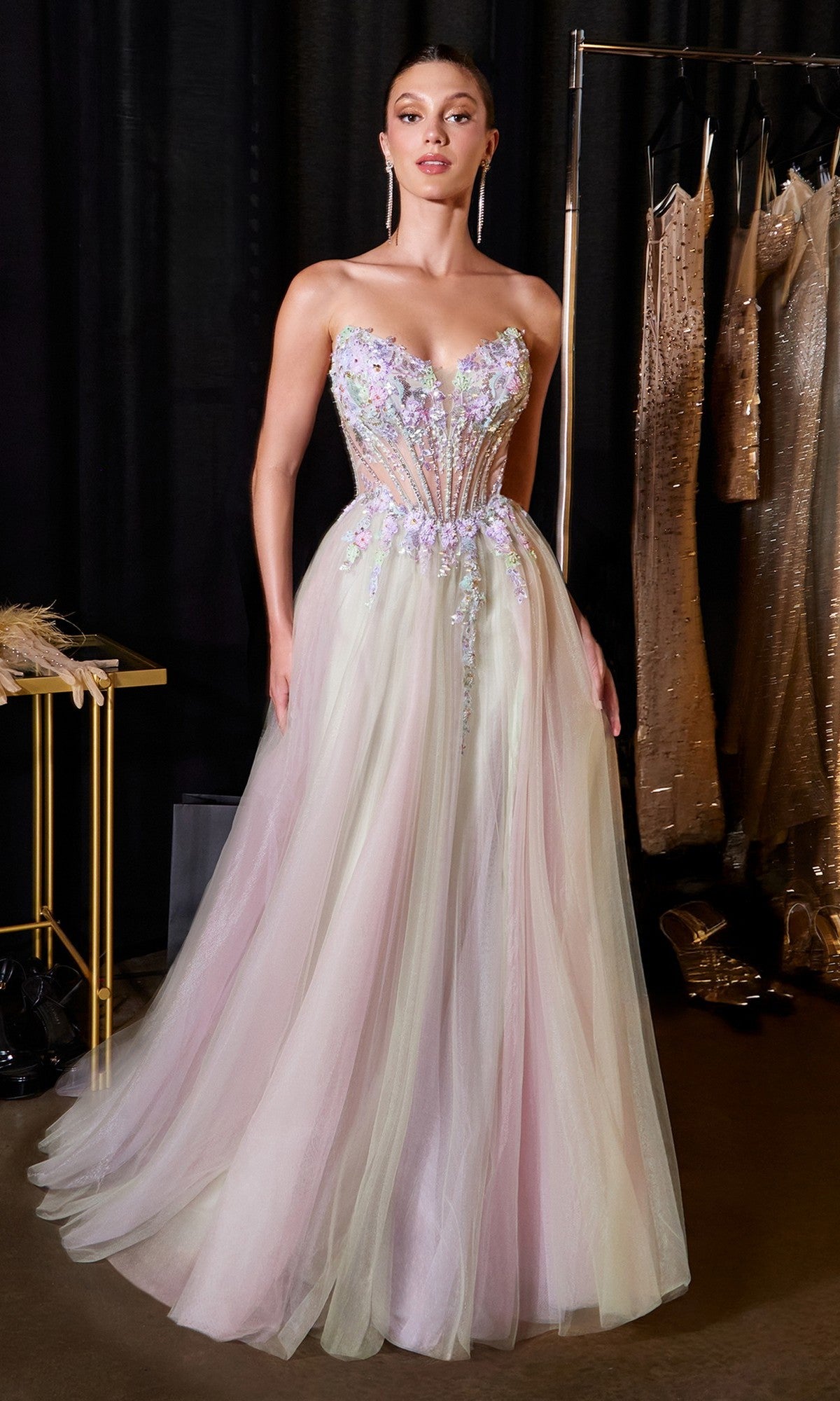 A woman wears the Ladivine CD2418 Long Prom Dress, featuring a strapless embroidered corset bodice and pastel tulle A-line skirt, standing amid hanging dresses and a table.